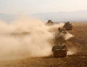 A convoy of land vehicles drive across a desert.